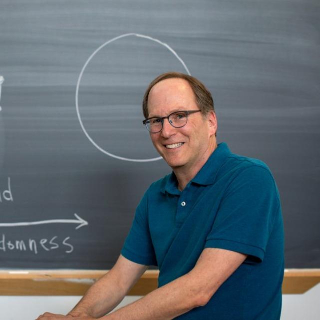 		Steven Strogatz in front of a blackboard with "small world" and an illustration on it showing a circle and interconnected lines inside
	